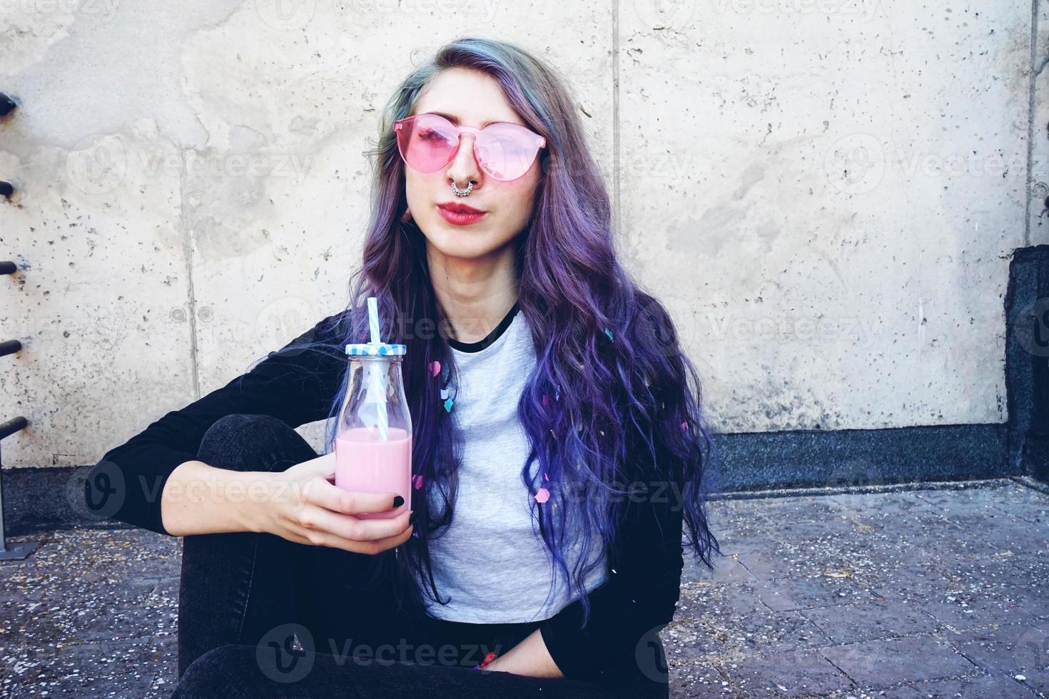 Feliz hermosa adolescente con gafas de sol rosa bebe y disfruta de una bebida rosa sentada en suelo urbano foto