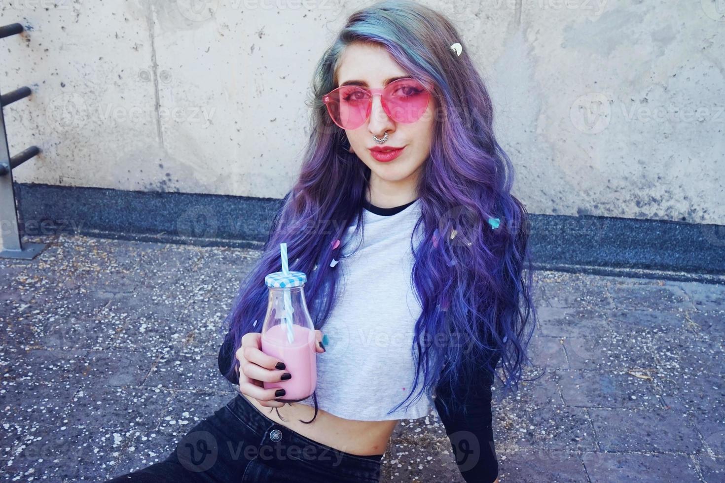 Happy beautiful teen with pink sunglasses drinks and enjoys a pink beverage sitting on urban ground photo