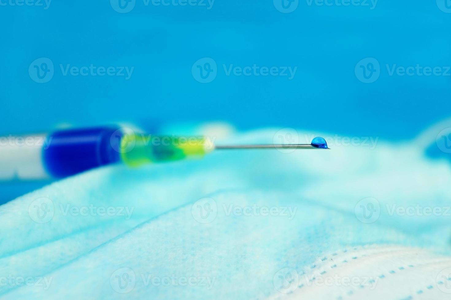 A detailed macro of a syringe with a blue liquid drop through the needle on a medical mask with blue background photo