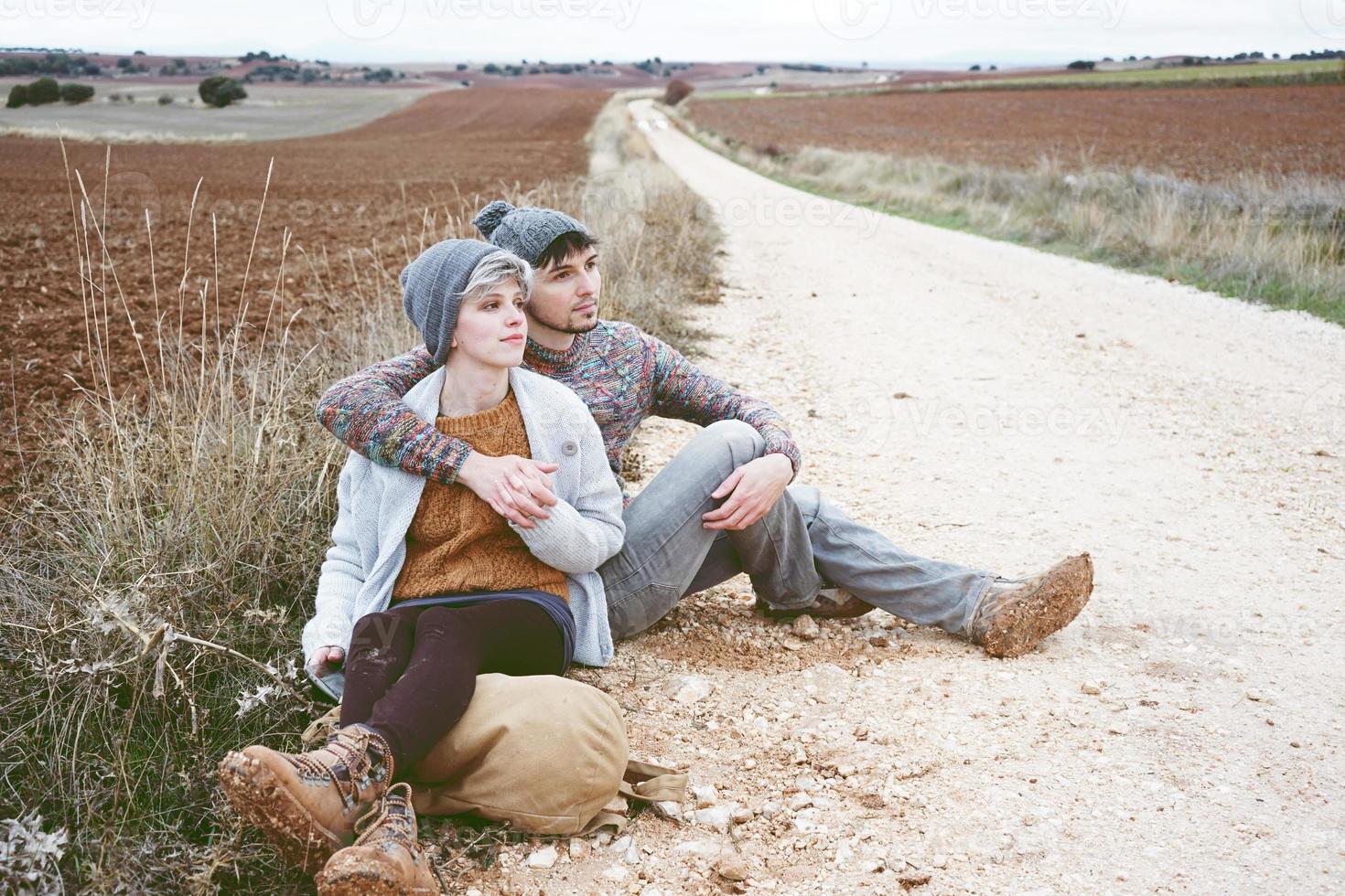 Pareja de jóvenes millennials abrazándose y descansando junto a una mochila en un viaje de aventura en un camino rural al aire libre foto