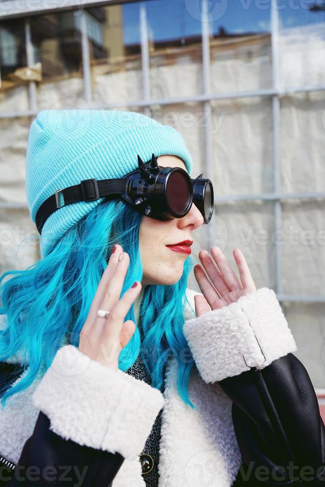 Retrato de una mujer joven punk o gótica con cabello de color azul y vistiendo gafas steampunk negras y gorro de lana azul en una calle urbana al aire libre foto