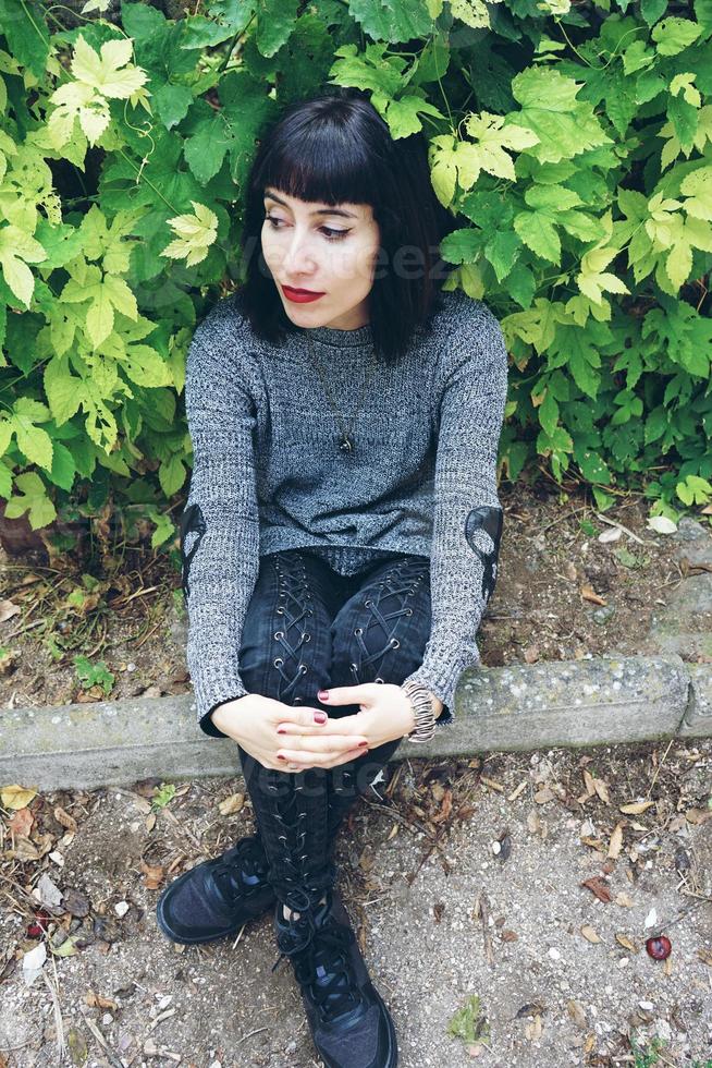 Beautiful caucasian brunette woman sitting on ground in a park wearing punk or gothic clothes and surrounded by green leafs photo