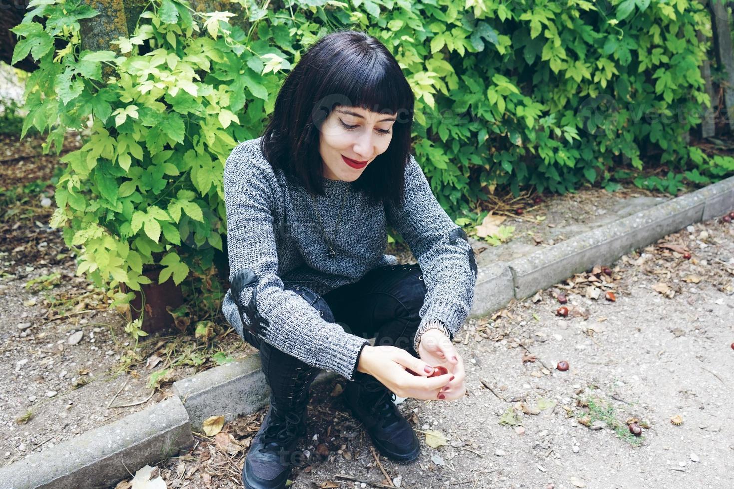 Beautiful caucasian brunette woman sitting on ground in a park wearing punk or gothic clothes and surrounded by green leafs photo