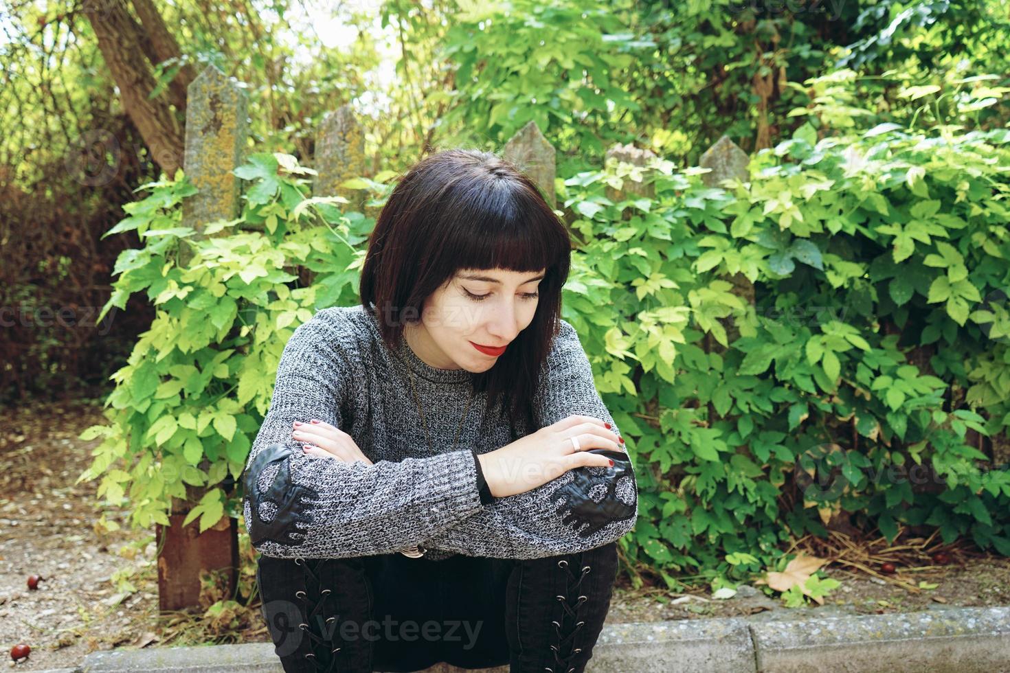 Beautiful caucasian brunette woman sitting on ground in a park wearing punk or gothic clothes and surrounded by green leafs photo