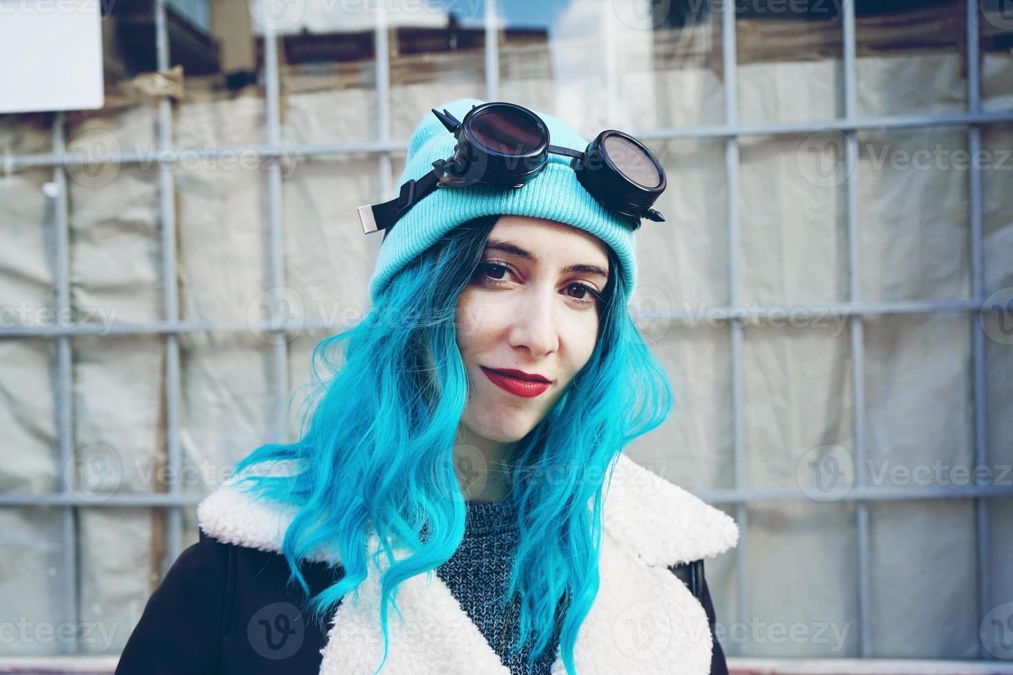 Portrait of a punk or gothic young woman with blue colored hair and wearing black steampunk glasses and blue wool cap in a urban outdoor street photo