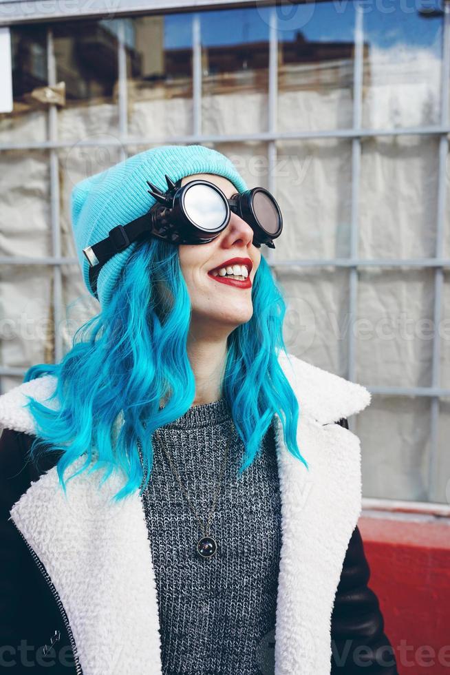 Retrato de una joven punk o gótica sonríe con el pelo de color azul y lleva gafas steampunk negras y gorro de lana azul en una calle urbana al aire libre foto