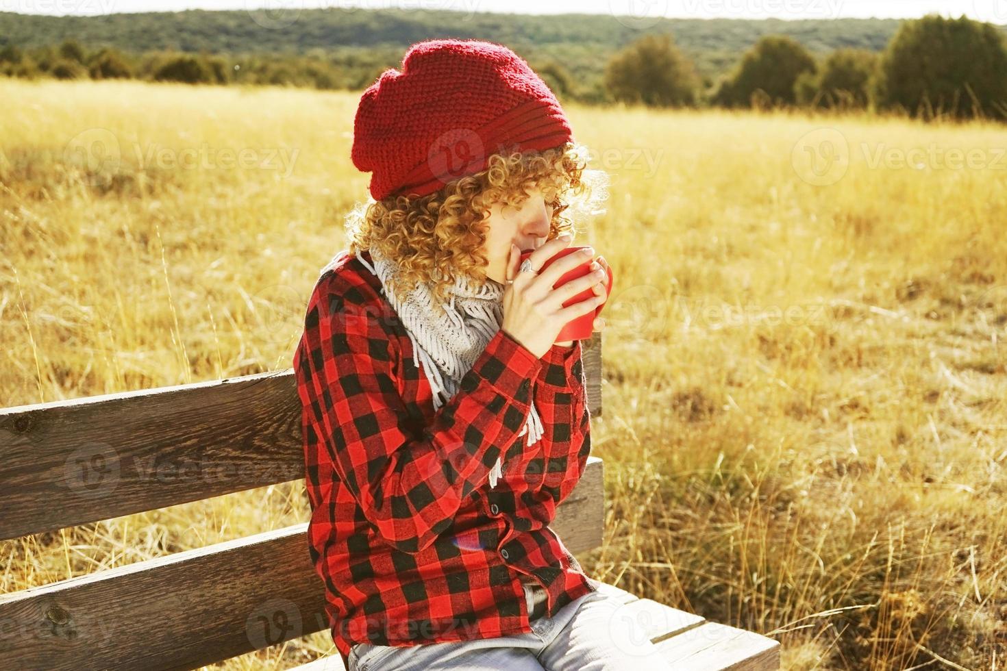 Retrato frontal de una mujer joven con camisa a cuadros roja con gorro de lana y bufanda tomando una taza de té o café mientras toma el sol sentada en un banco de madera en un campo amarillo con luz de fondo del sol otoñal foto