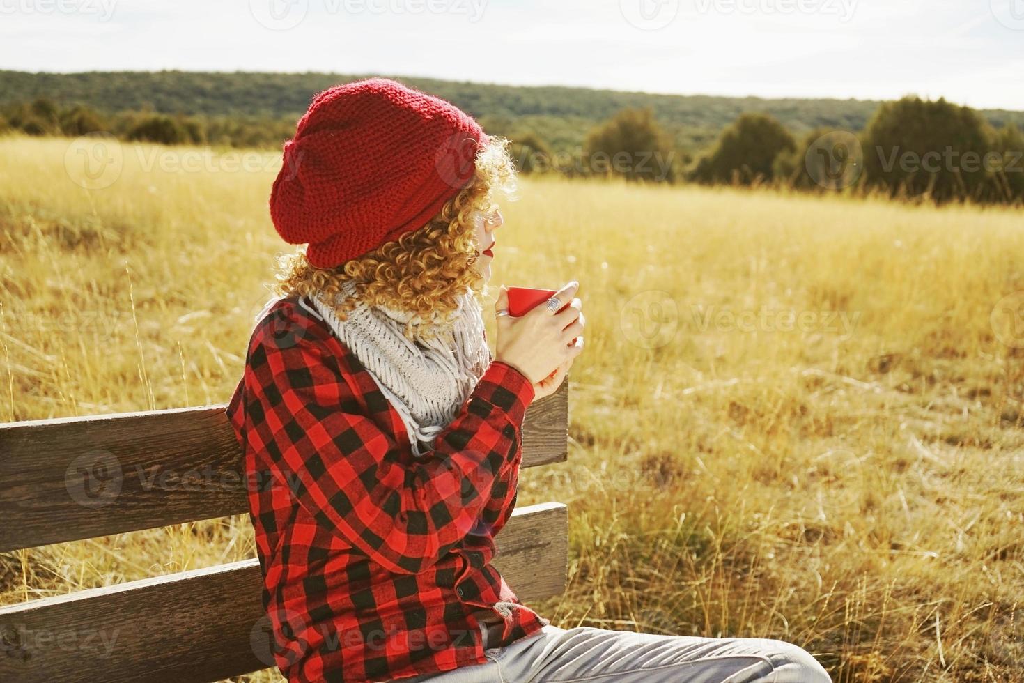 A young woman from behind in red plaid shirt with a wool cap and scarf taking a cup of tea or coffee while she is sunbathing sitting in a wooden bench in a yellow field with backlight from autumn sun photo