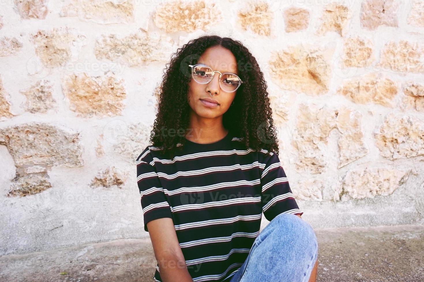 Un retrato de mujer negra joven seria con gafas, jeans y una camiseta a rayas, sentada en el suelo y cabello rizado foto