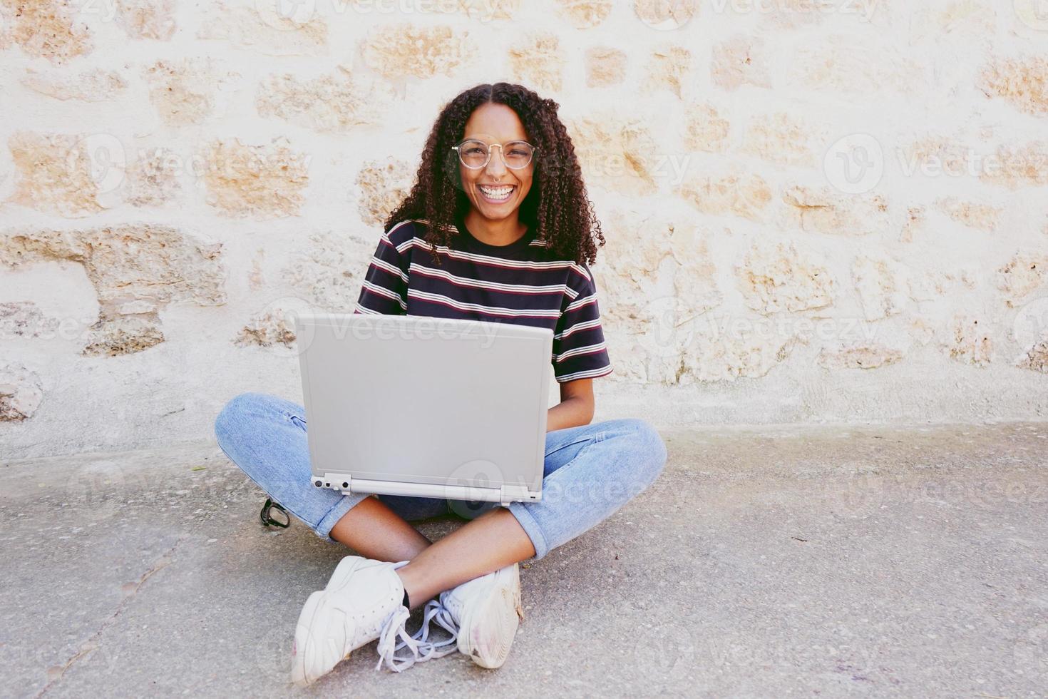 Un retrato de una mujer negra joven y sonriente feliz con el pelo rizado con gafas, jeans y una camiseta a rayas, sentada en el suelo y trabajando o haciendo la tarea foto