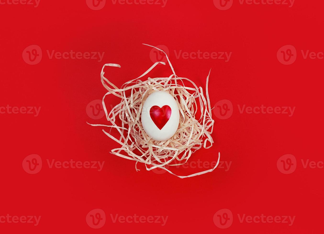 White egg with heart shape in decorative nest on red background. photo