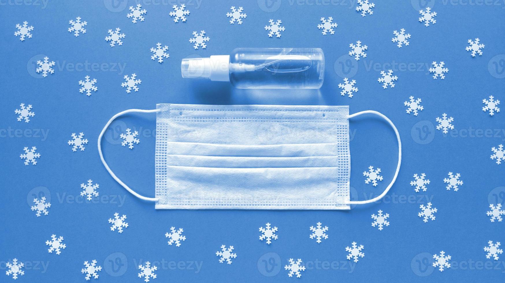 White medical mask and hand sanitizer in transparent bottle with spray cap at the middle of blue background and scattered white snowflakes on it. Simple festive flat lay. Medical concept. Stock photo. photo