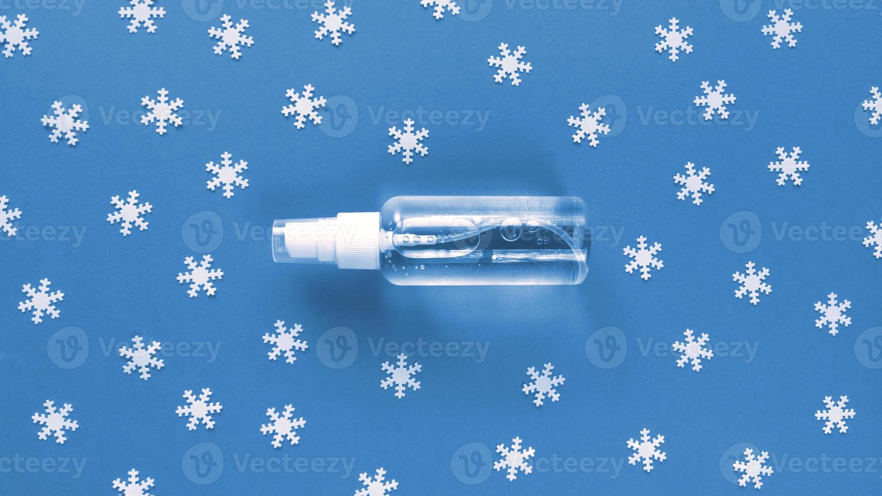 Hand sanitizer transparent bottle with spray cap at the middle of blue background and scattered white snowflakes on it. Simple festive flat lay with pastel paper texture. Medical concept. Stock photo. photo