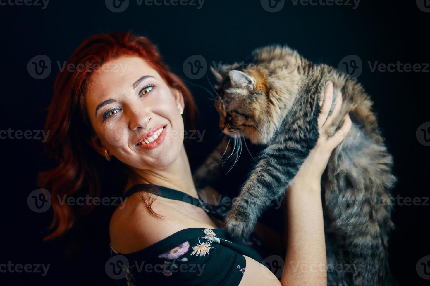 mujer pelirroja abraza gato esponjoso sobre fondo negro. foto
