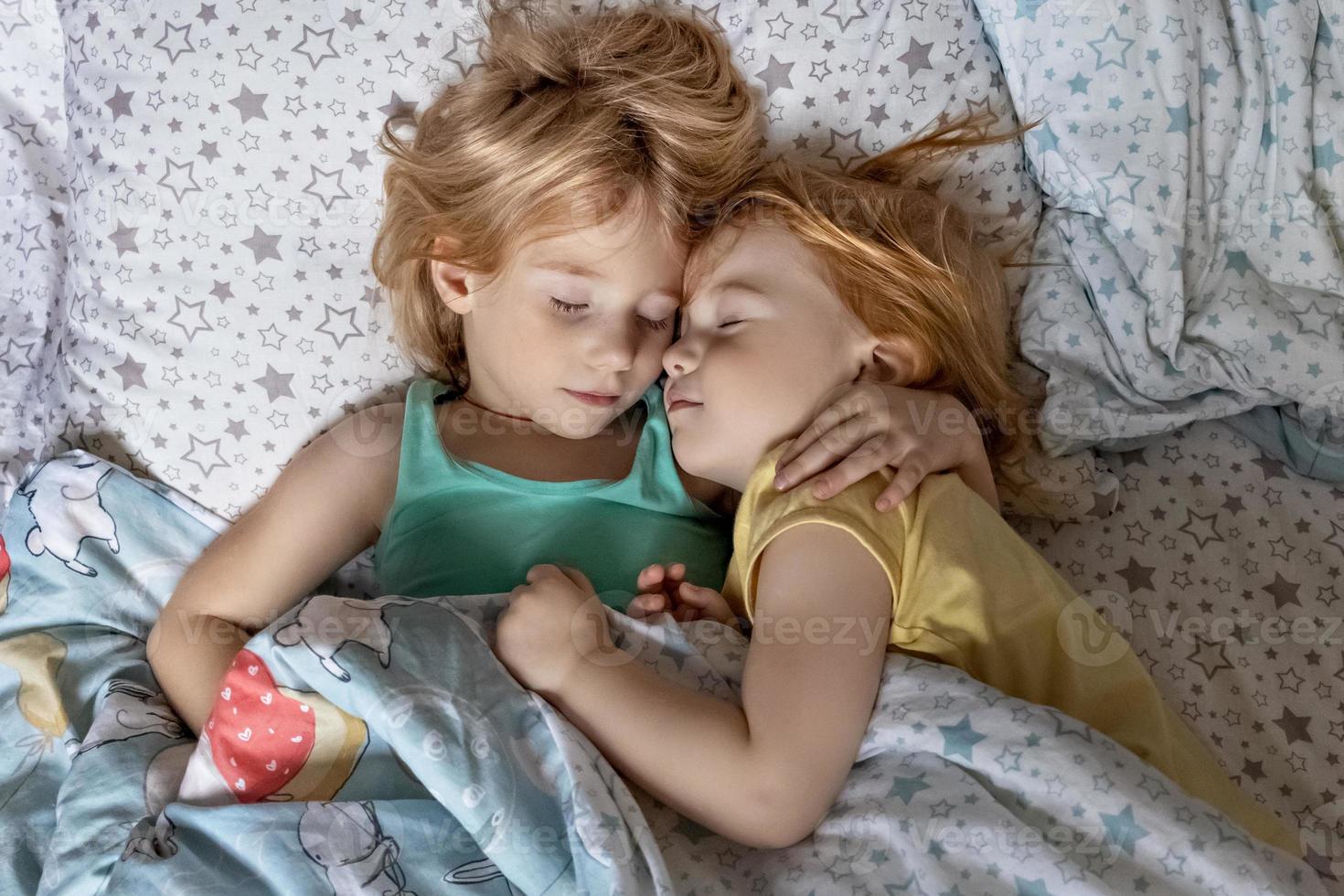 Two little sibling girls sisters sleeping in an embrace in bed under one blanket photo