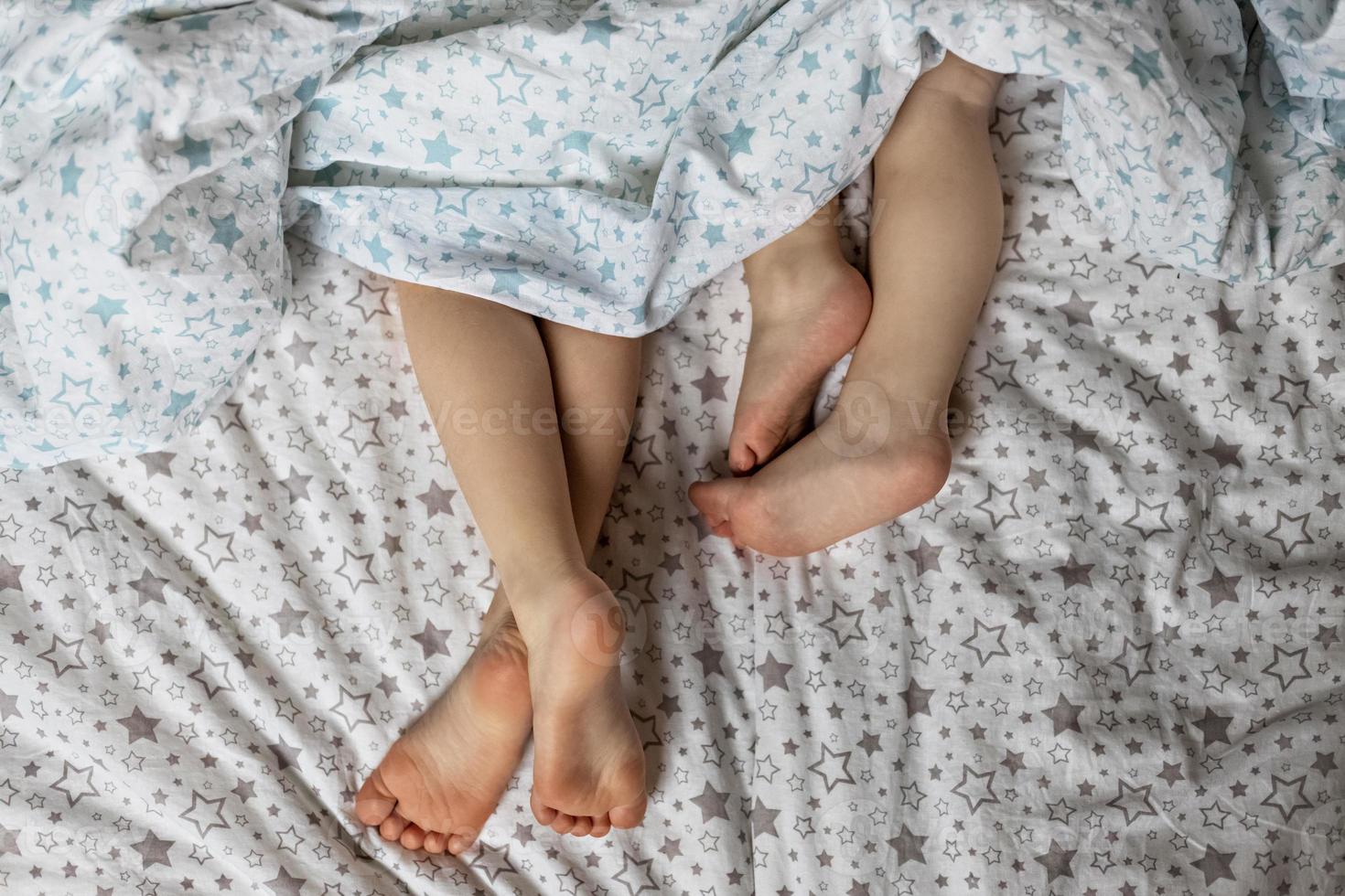 Close-up of two pairs of toddler girls feet on the bed under the blanket. Light blue and beige tones. photo