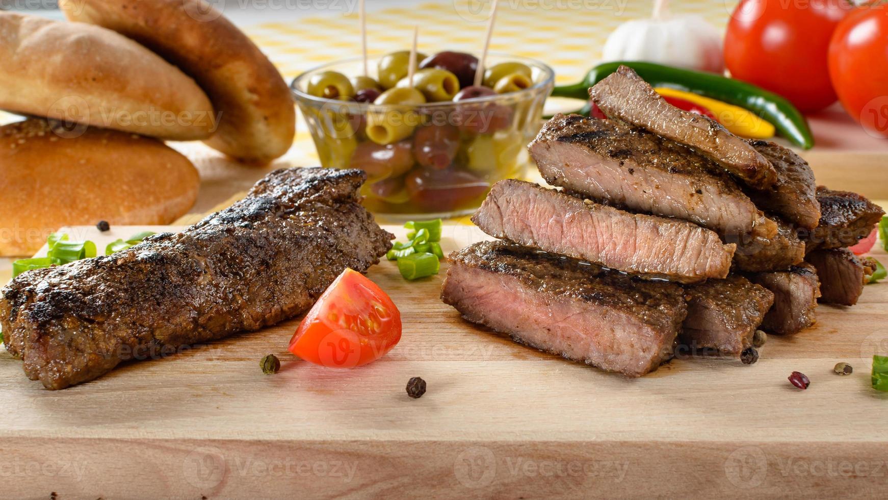 Grilled beef meat on wooden plate with vegetables and olives in the background. photo