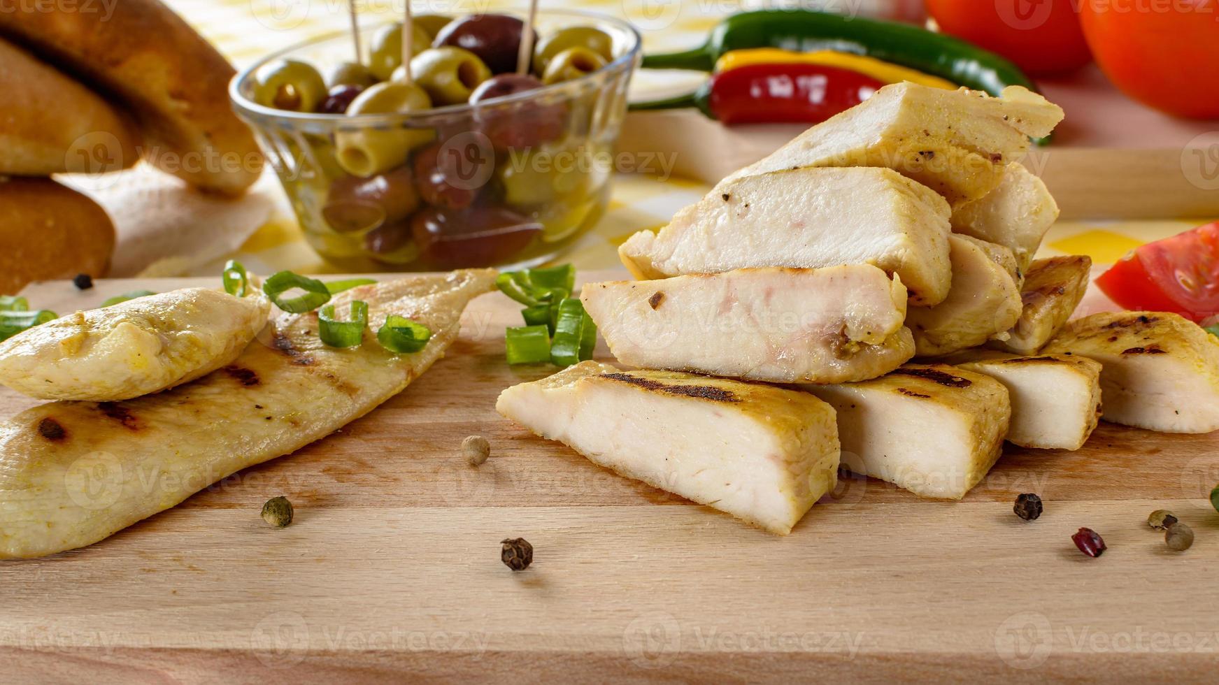 Grilled chicken meat on wooden plate with vegetables and olives in the background. photo
