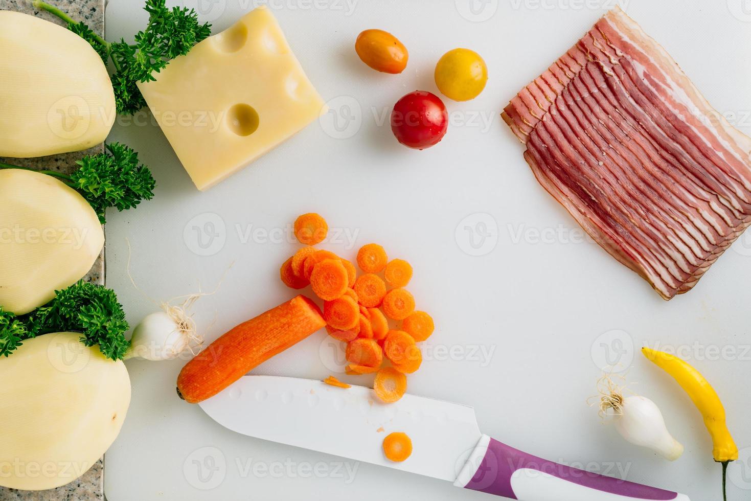 Sliced carrot with knife, potato, cheese, bacon, some onion and herbs on table. photo