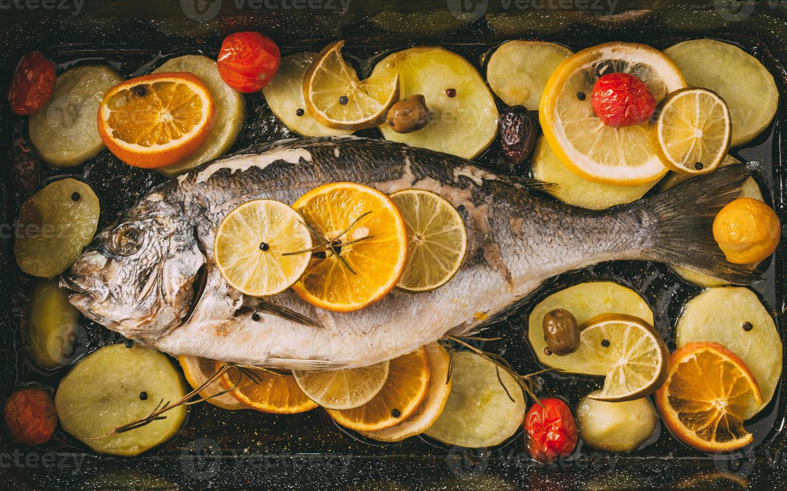 Sea gilt-head bream fish in the baking tray baked with potatoes, rosemary, lemon, orange, olives, tomatoes, onion and lime. Fresh Orata, Dorade fish preparation. photo