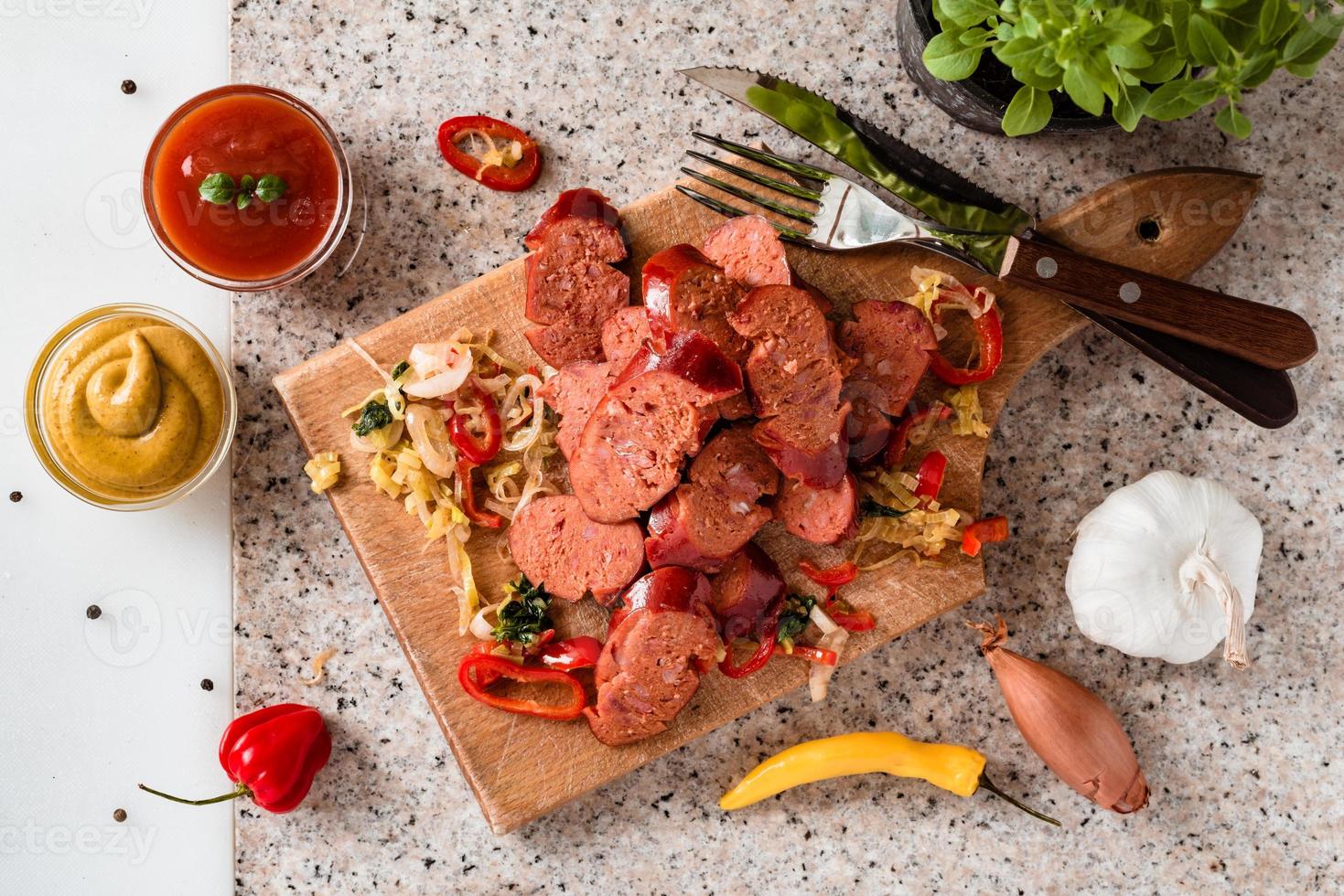 Sliced sausage and mix of fresh vegetables served on a rustic wooden board, mustard, ketchup, fresh hot chili peppers and herbs. photo