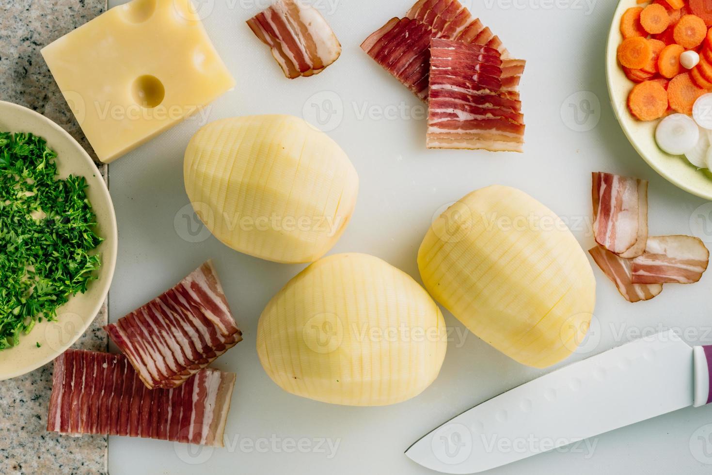 Sliced potato and bacon with knife,potato ready to be filled with bacon. Cheese, carrot some onion and herbs on table. photo