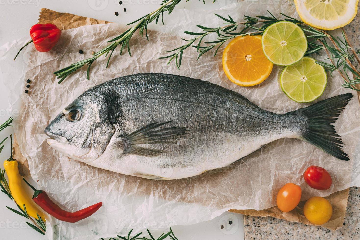 Sea gilt-head bream fish on baking sheet with rosemary, lemon, orange, tomato, hot peppers and lime. Fresh Orata, Dorade fish on kitchen table. photo