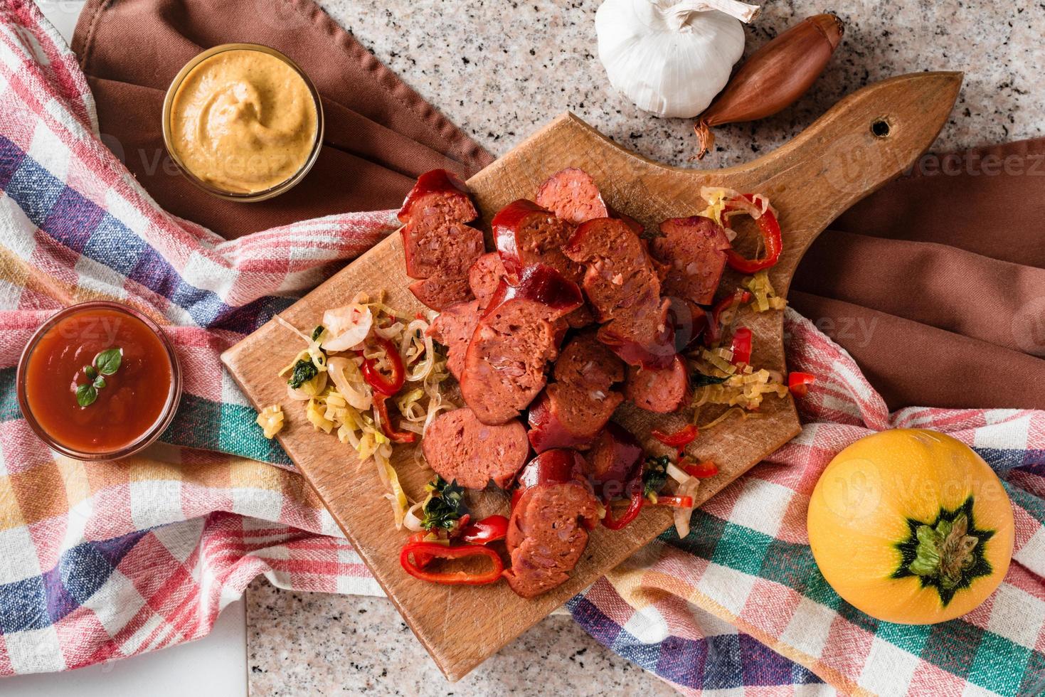 Sliced sausage and mix of fresh vegetables served on a rustic wooden board, mustard, ketchup, fresh hot chili peppers and herbs. photo
