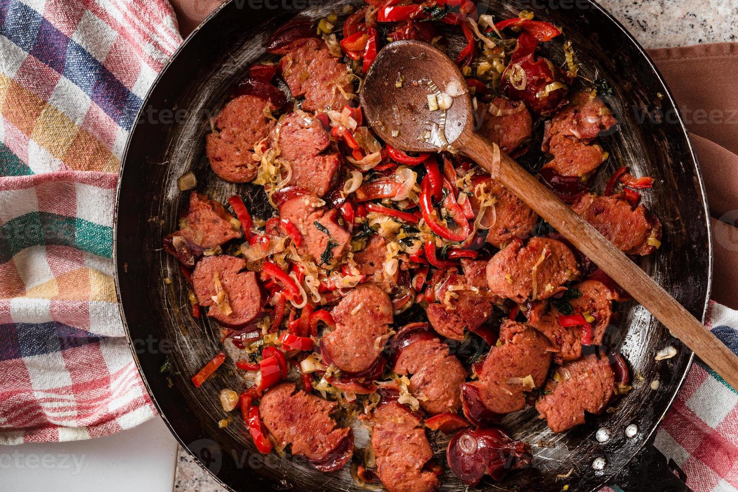 Sliced sausage and mix of fresh vegetables fried in the rustic pan with scoop on dishcloth. photo