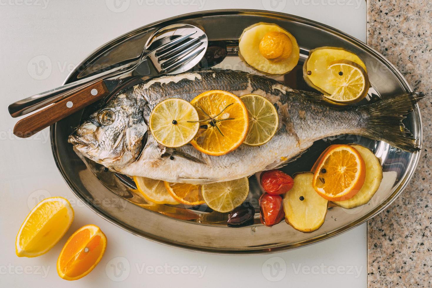 Sea gilt-head bream fish on the plate baked with potatoes, rosemary, lemon, orange, olives, tomatoes and lime. Fresh Orata, Dorade fish preparation. photo