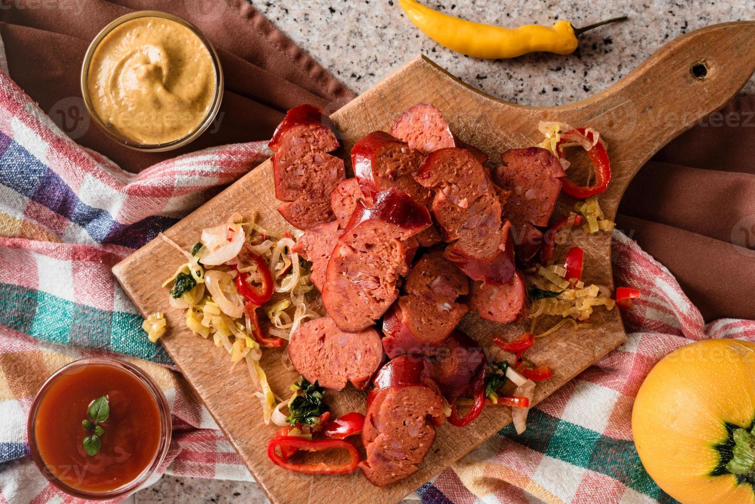 Sliced sausage and mix of fresh vegetables served on a rustic wooden board, mustard, ketchup, fresh hot chili peppers and herbs. photo