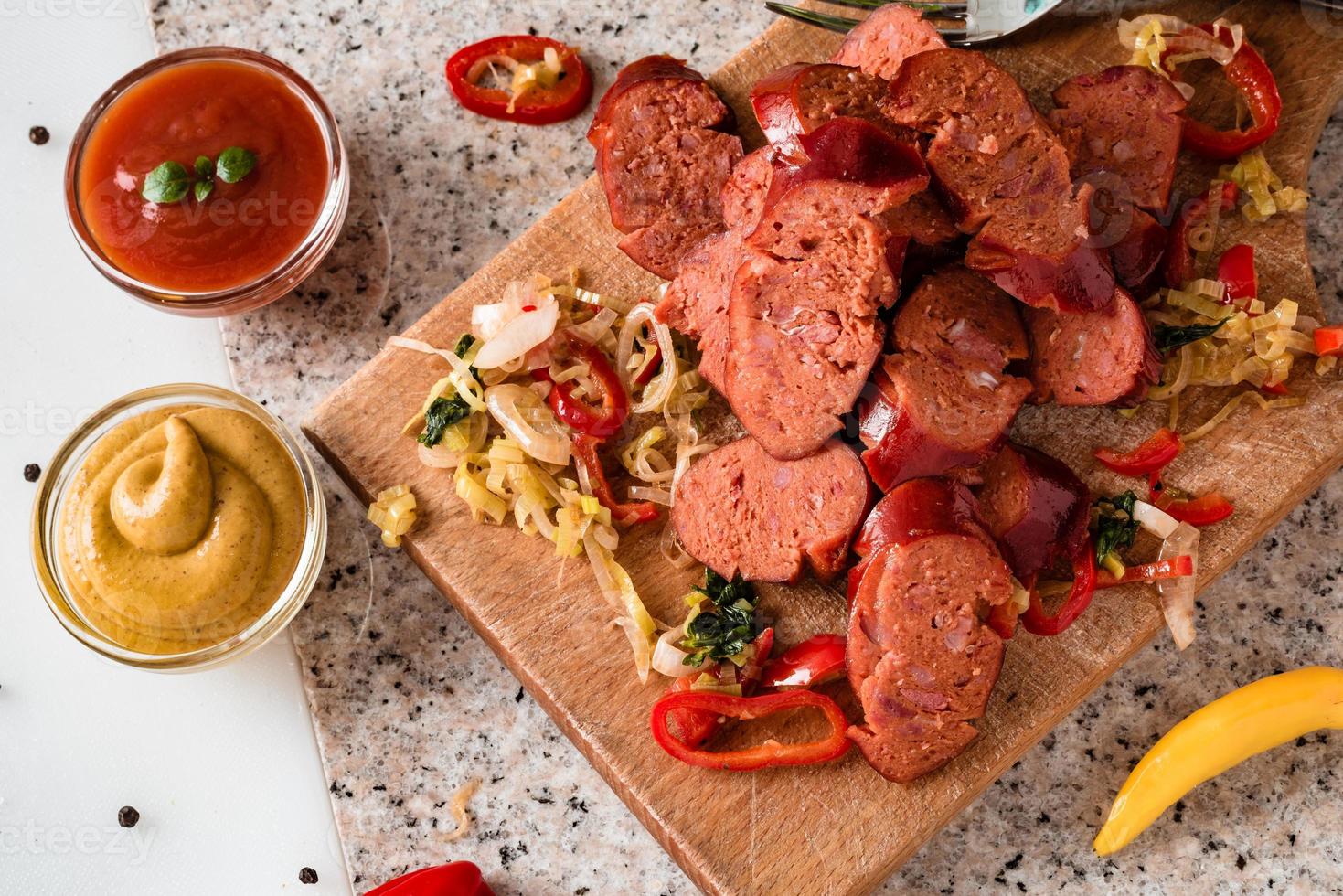 Sliced sausage and mix of fresh vegetables served on a rustic wooden board, mustard, ketchup, fresh hot chili peppers and herbs. photo