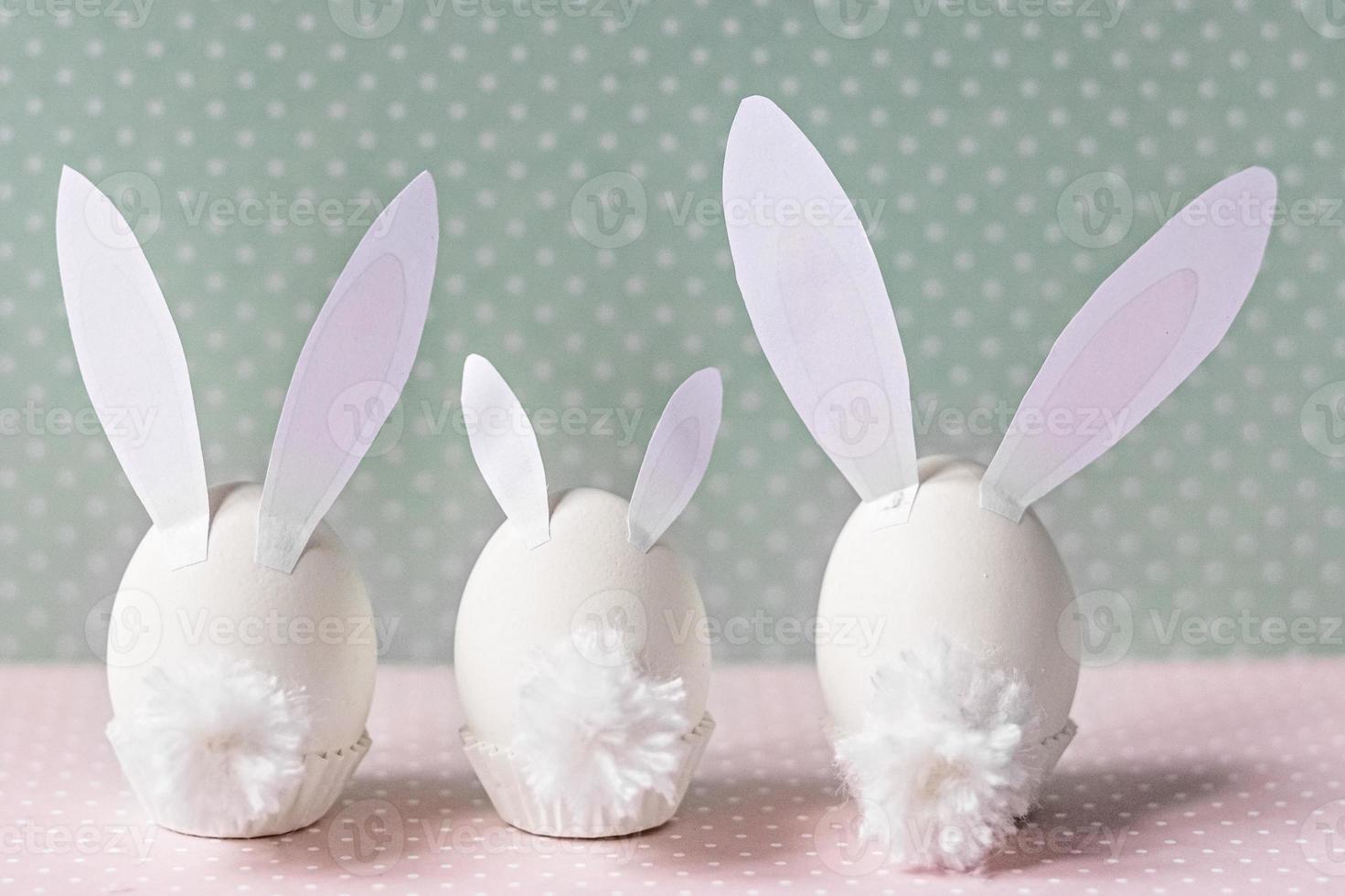 White chicken eggs with bunny ears and tails on bed flowers background. A family. Happy Easter holiday concept photo