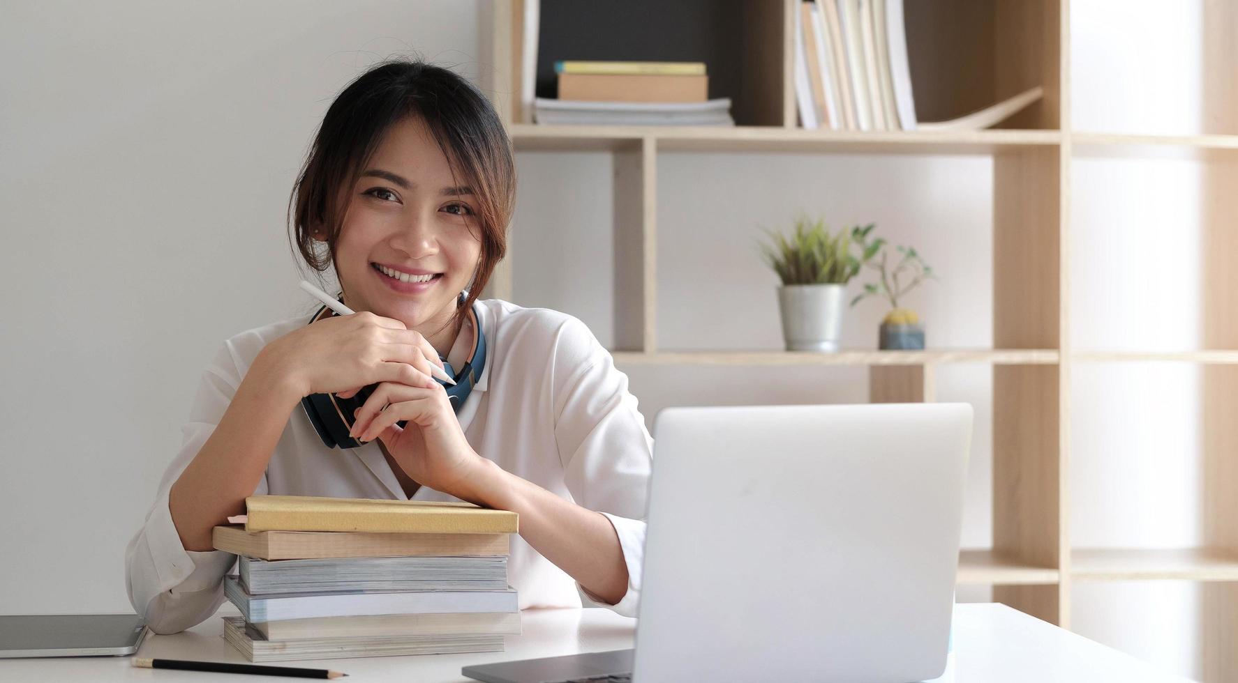 Sonriente mujer asiática que trabaja en el escritorio con libros y computadora portátil foto