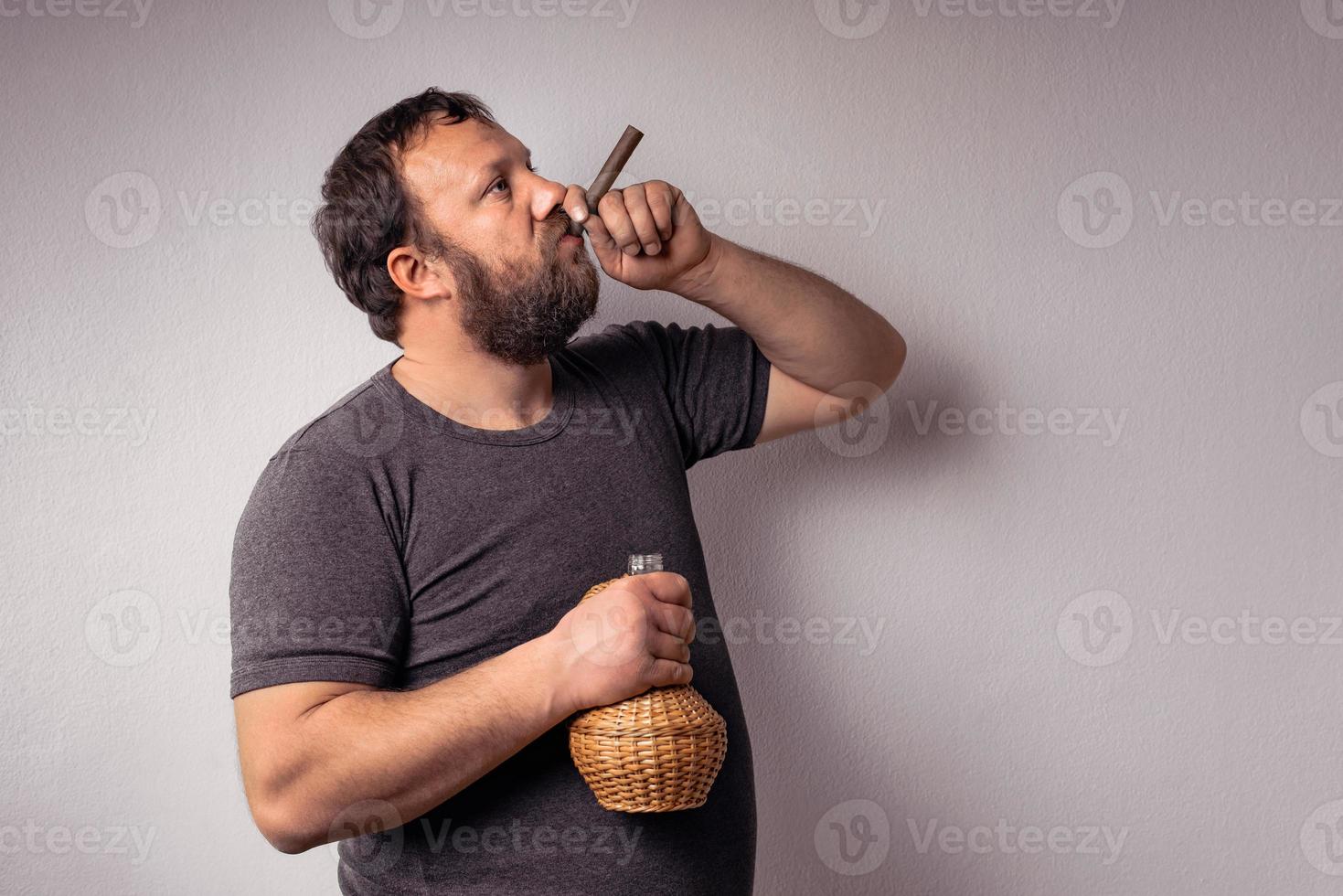 Hombre barbudo sostiene una botella de alcohol y fuma su cigarro y celebra foto