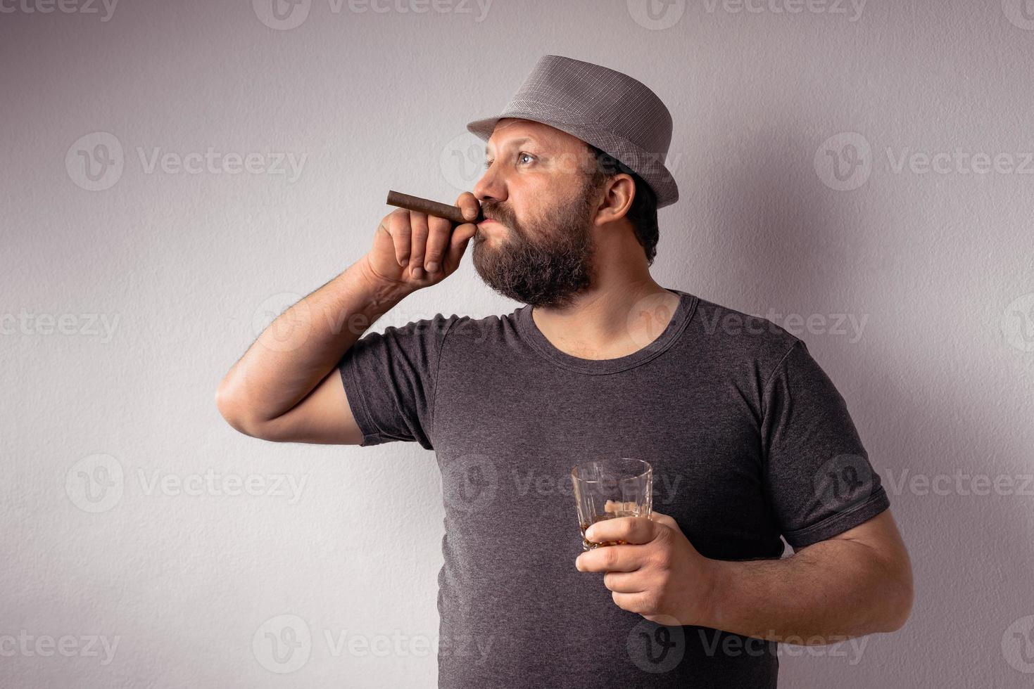apuesto hombre barbudo con camiseta gris y sombrero bebiendo un licor fuerte foto