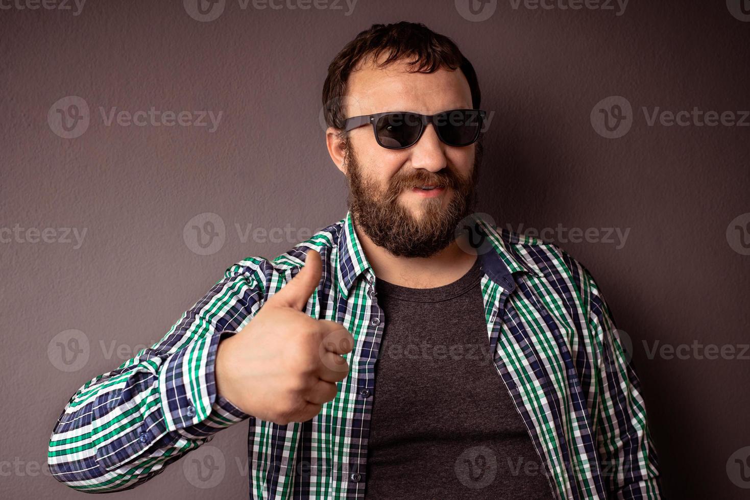 apuesto hombre barbudo hipster con gafas de sol y camisa mostrando el pulgar hacia arriba foto