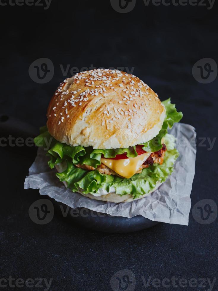 apetitosa hamburguesa casera fresca, con chuleta de pollo, lechuga, tomate, queso y salsa. sobre una tabla de madera sobre un fondo negro foto
