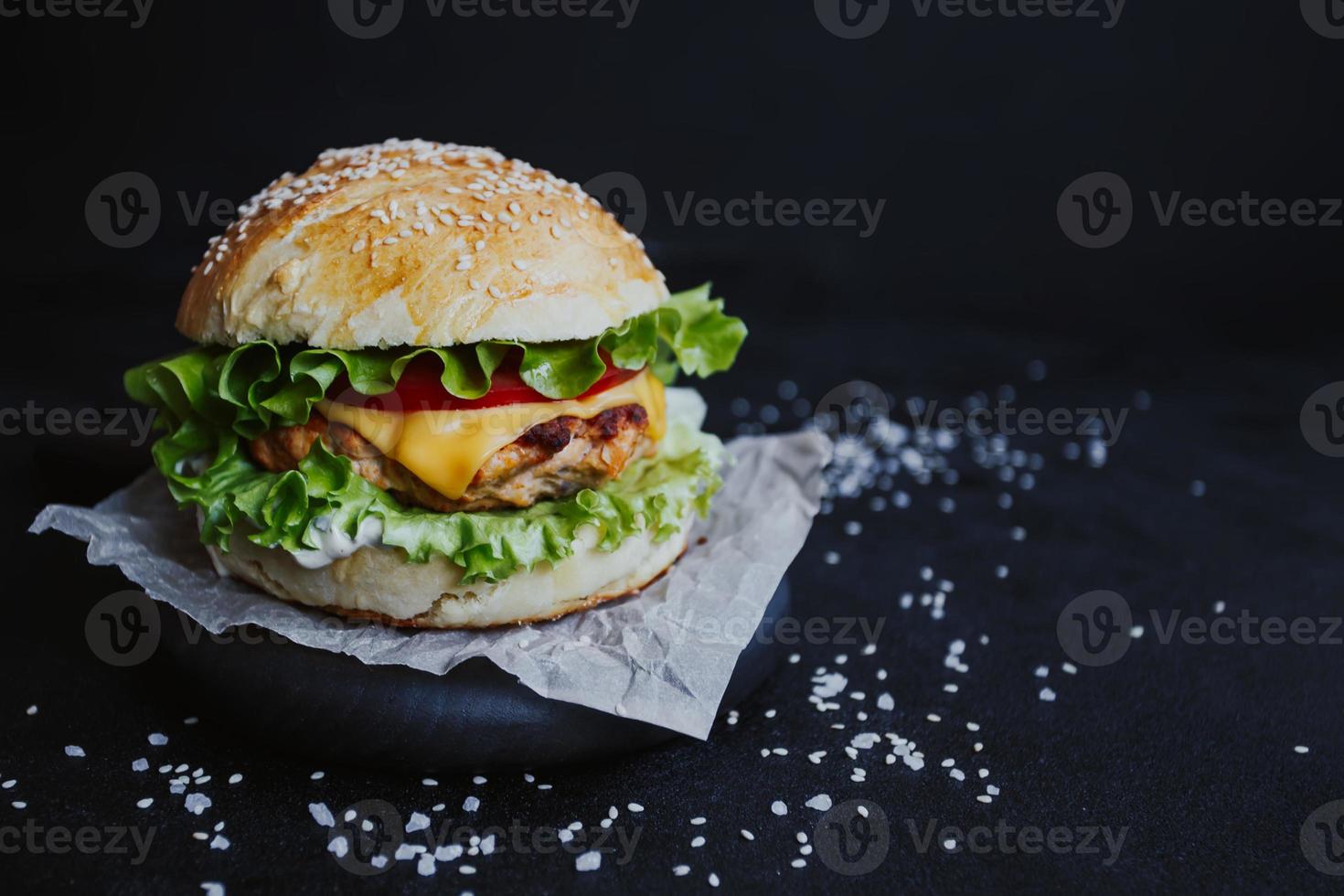 Appetizing fresh homemade burger, with chicken cutlet, lettuce, tomatoes, cheese and sauce. On a wooden board on a black background photo