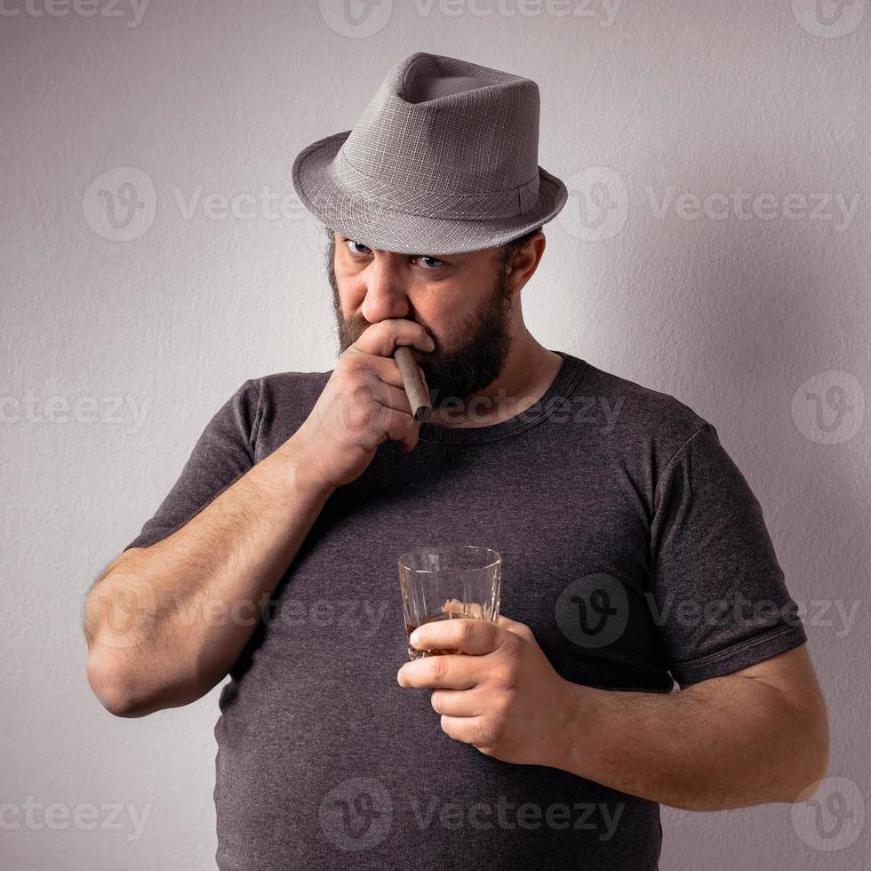 apuesto hombre barbudo con camiseta gris y sombrero bebiendo un licor fuerte foto