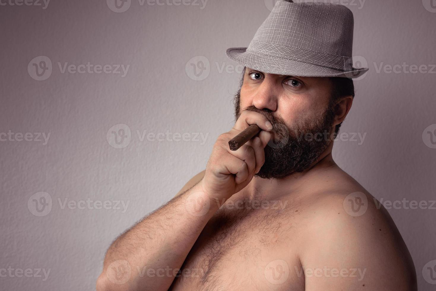 Apuesto hombre barbudo semidesnudo con sombrero gris fumando su cigarro y de pie contra la pared de fondo gris. foto