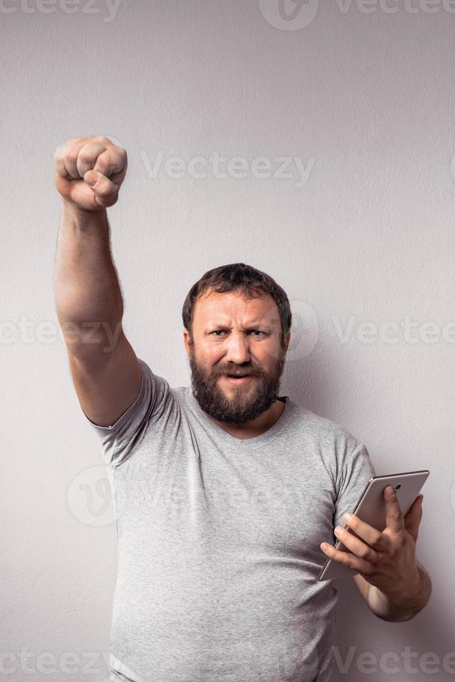 Happy handsome bearded excited man in gray t-shirt with hand up holding his tablet and celebrating photo