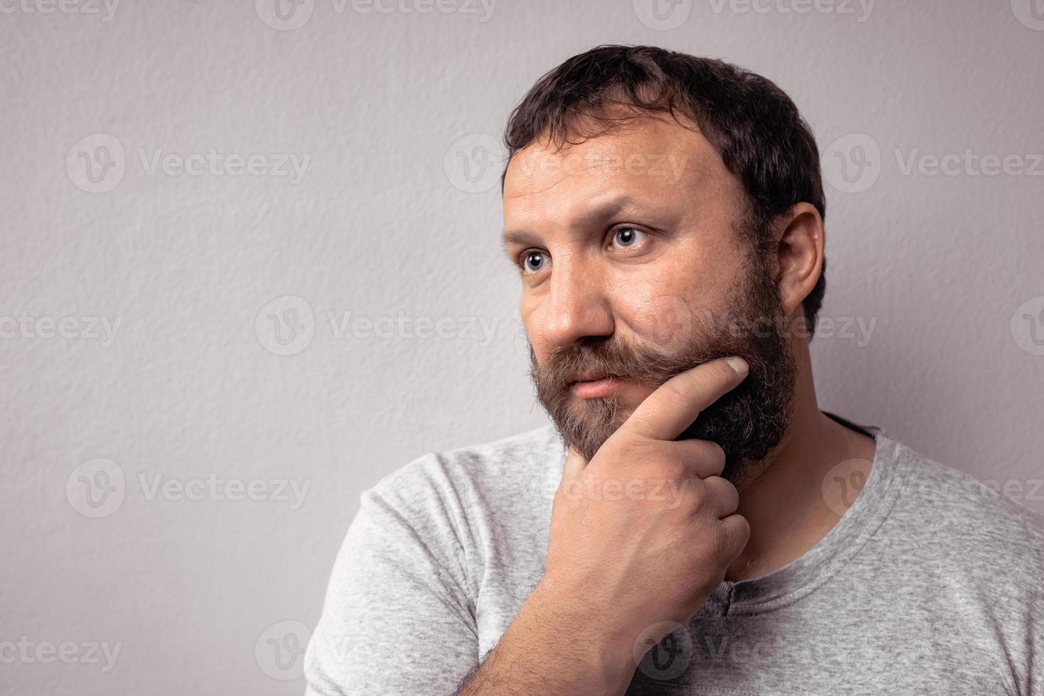 Bearded man in gray t-shirt looking away while keeping his hand over his beard photo