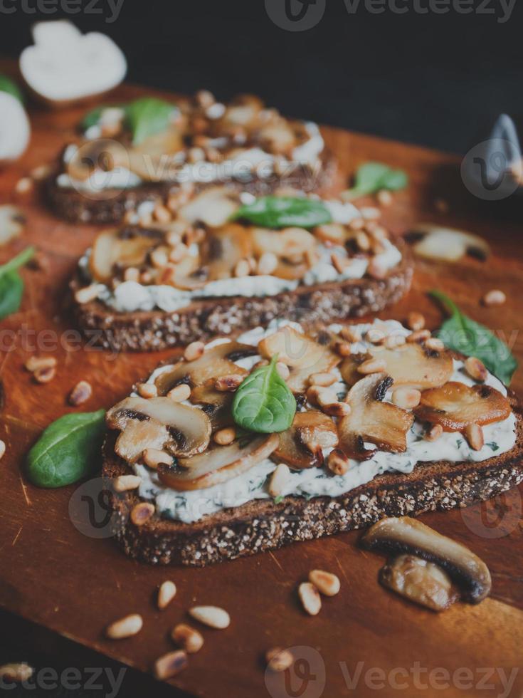 Tasty fresh bruschetta with mushrooms, spinach, garlic, cream cheese and pine nuts, on a wooden board, on a dark background. photo