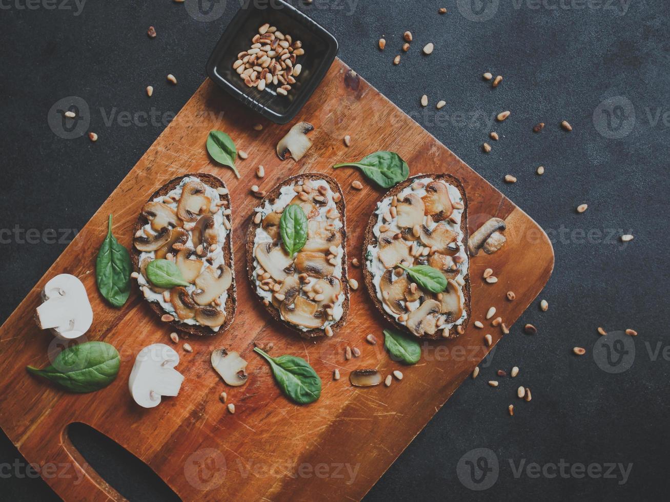 Sabrosa bruschetta fresca con champiñones, espinacas, ajo, queso crema y piñones, sobre una tabla de madera, sobre un fondo oscuro. foto