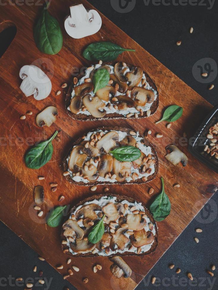 Sabrosa bruschetta fresca con champiñones, espinacas, ajo, queso crema y piñones, sobre una tabla de madera, sobre un fondo oscuro. foto