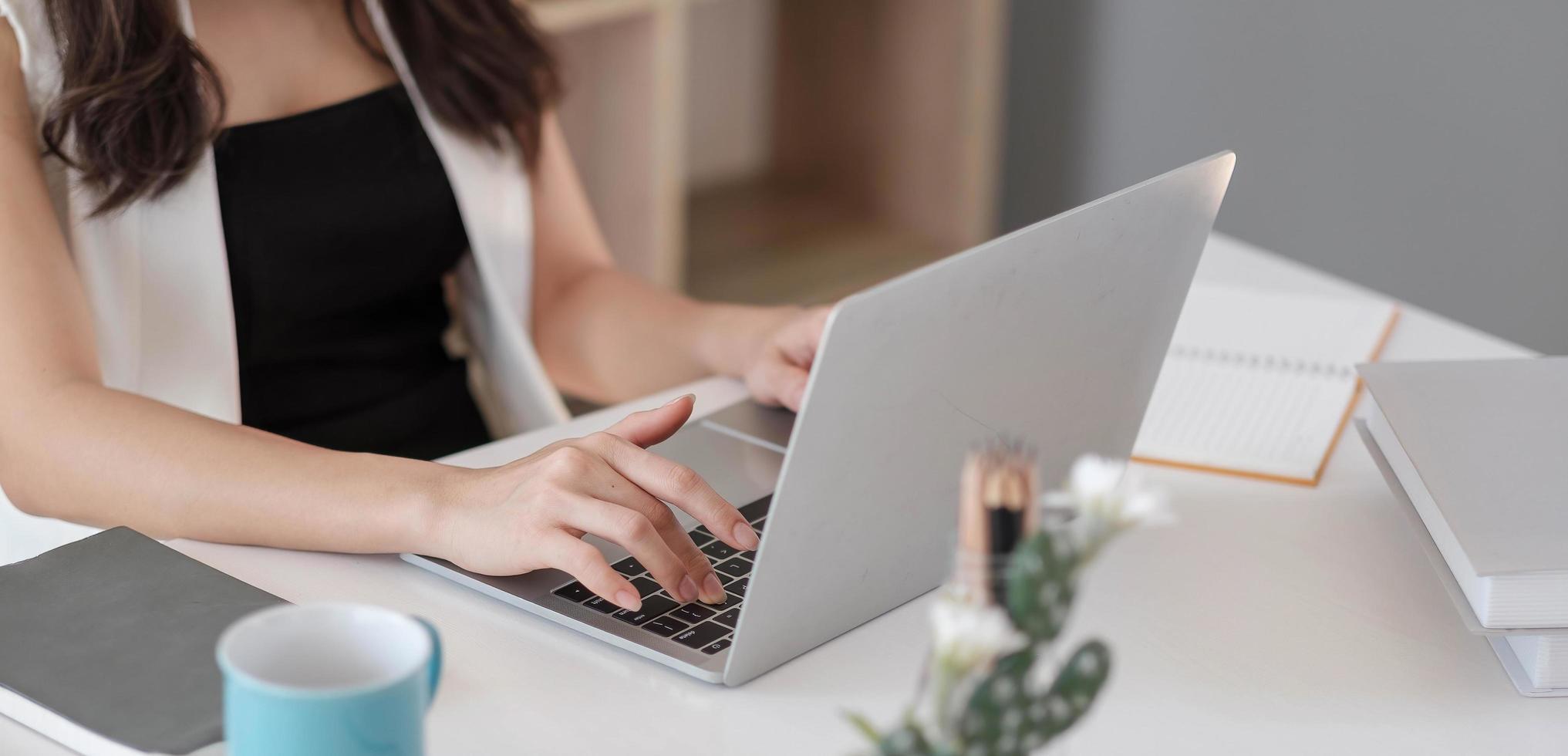 manos de empresaria escribiendo, navegando por internet en el teclado de la computadora portátil con teléfono inteligente móvil, auriculares, una taza de café en la mesa de oficina de madera en la oficina en casa. estudiante femenina aprendiendo en línea foto