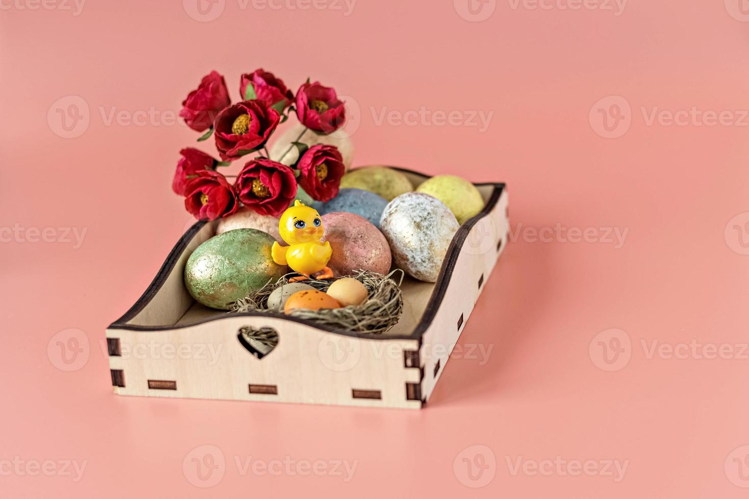 Huevos de pascua en un nido natural en una bandeja de madera, flores y un pollo decorativo sobre un fondo rosa. foto