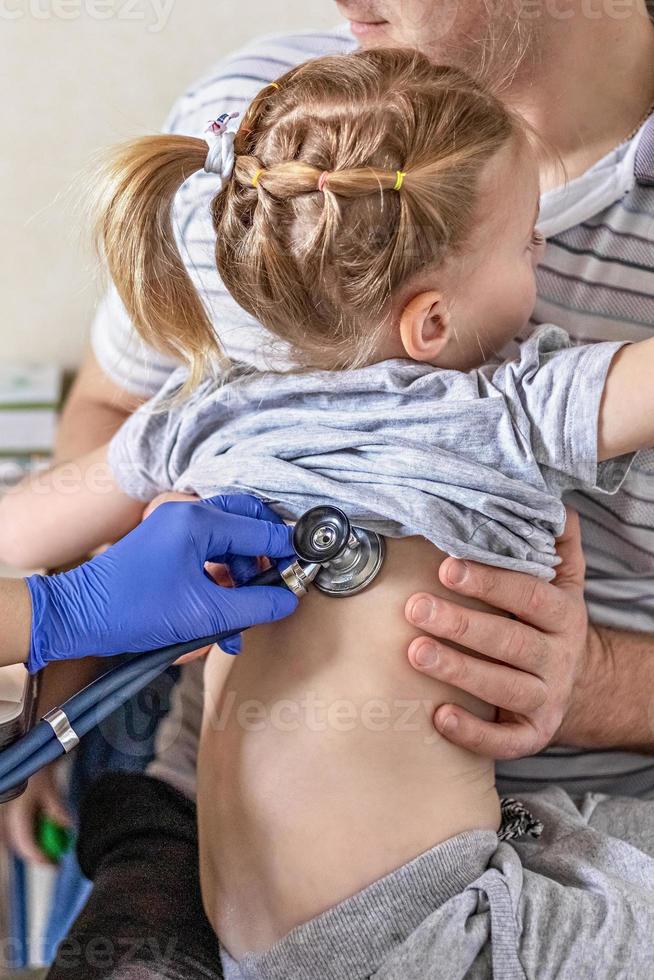 niña en los brazos de su padre en el consultorio del médico en la clínica. el médico examina al niño, escucha los pulmones con un fonendoscopio. tratamiento y prevención de infecciones respiratorias. foto