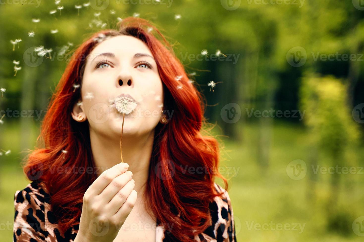 Woman with red hairs blows away a dandelion in the park photo